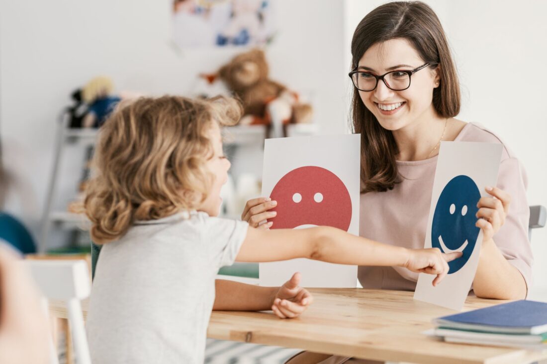 White blonde haired child choosing smiley face card from choice of two options white smiling woman with brown hair and glasses if offering her from across a table.