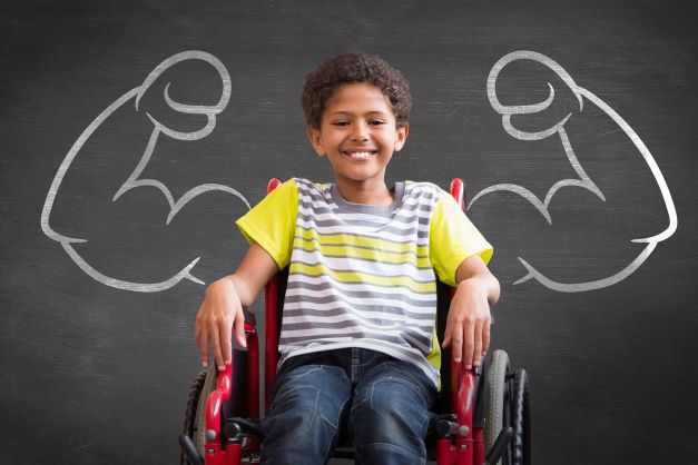 Black boy smiling, sat in wheelchair with picture of strong arms chalked onto blackboard behind him on wall