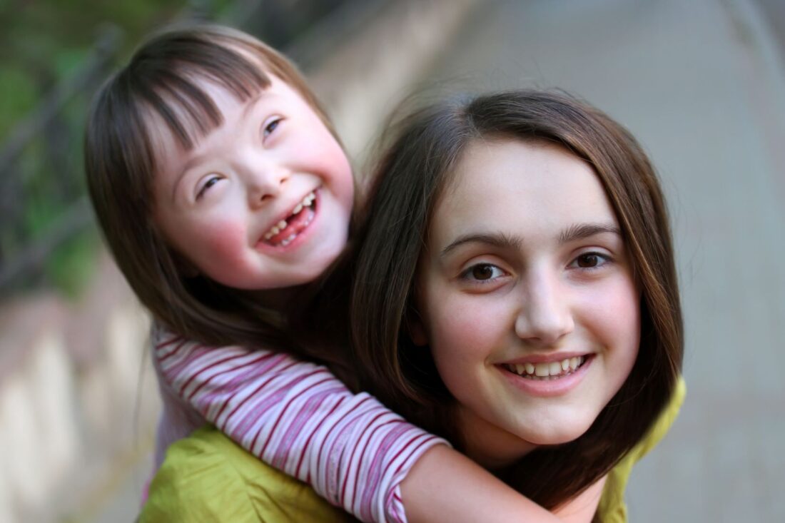 Smiling white girl with Down Syndrome with borwn hair and a pink stripy top. She is being carried on the back of her older sister a white non disabled teen with brown hair and wearing a yellow top