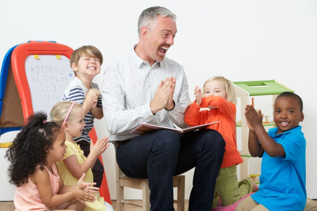 While man with grey hair, wearing a white shirt and navy trousers sitting down smiling and clapping his hands whilst storytelling to a group of young children