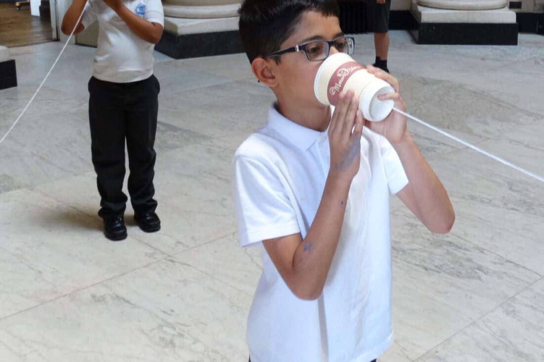 Schoolboy with dark short hear wearing glasses, speaking into a cup on a string with another child.