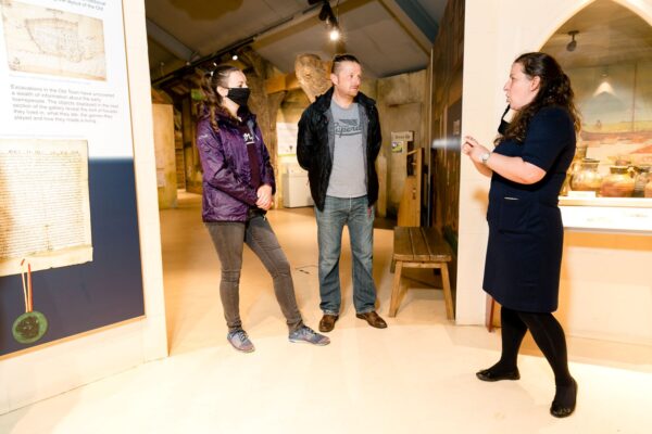 Image of three white adults (two women and a man) standing in a museum exhibition. One of the women is signing using BSL