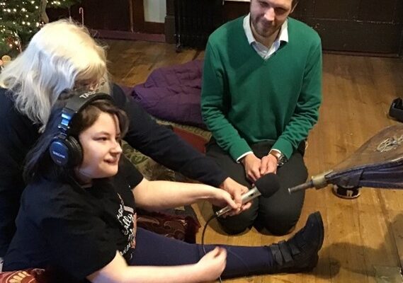 Yong white female wearing headphones ad dark top and trousers, sits on the floor legs outstretched. She hols a microphone in front of her pointing it to a pair of old bellows being opened by someone off screen. a white man wearing a green jumper sits near to ger watching and smiling. A white woman with white hair sits slightly behind her helping her to hold the microphone.