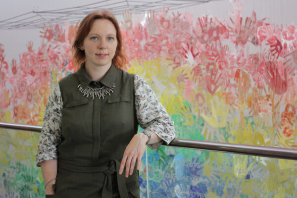 White woman with short red hair wearing a patterned top and waistcoat leans against a handrail with rainbow colours of hand prints on the wall behind her.